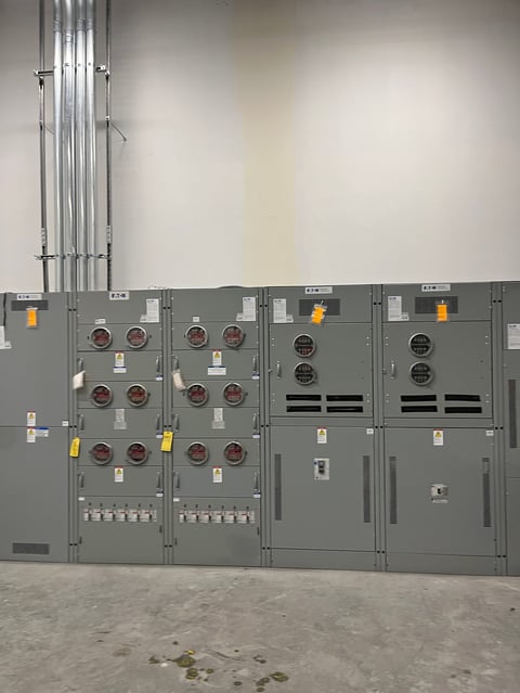 Tablero eléctrico industrial con gabinetes metálicos grises, medidores rojos, rejillas negras y tuberías conduit montadas en pared
