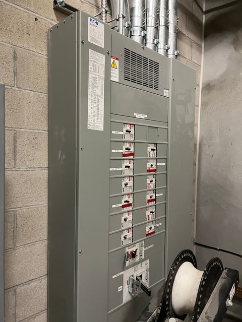 Industrial electrical panel mounted on concrete wall with circuit breakers, warning labels, and metal conduit pipes above