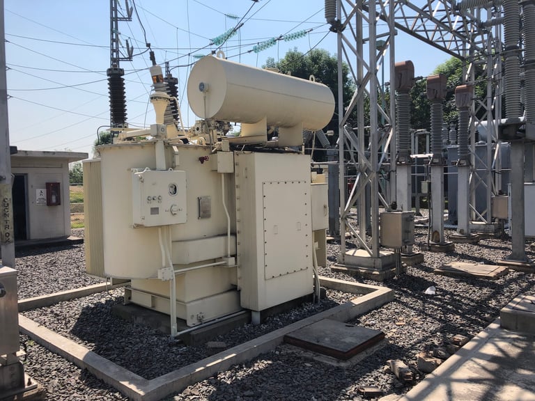 Large beige electrical transformer with cylindrical cooling tank mounted on concrete pad at industrial power substation with steel transmission towers and electrical equipment