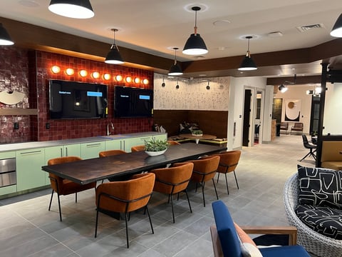 Modern office break room with dark wood dining table, orange chairs, mint green cabinetry, and red brick wall with illuminated signage