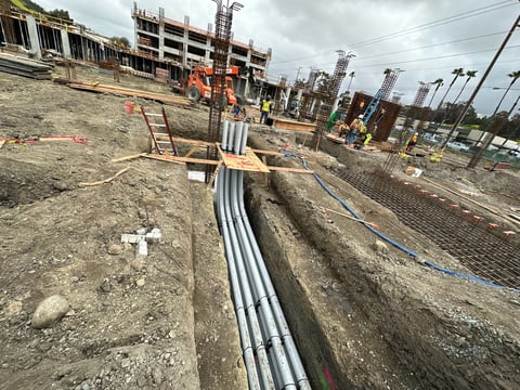 Construction site with metal pipes running through trenches, partially completed concrete structures in background, and construction workers and equipment visible
