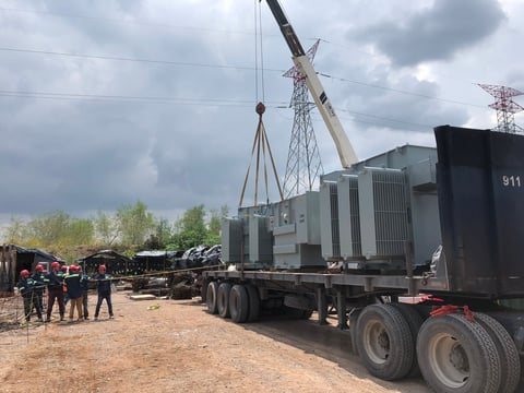 Large transformer equipment being lifted by crane onto truck at industrial construction site with power transmission towers in background