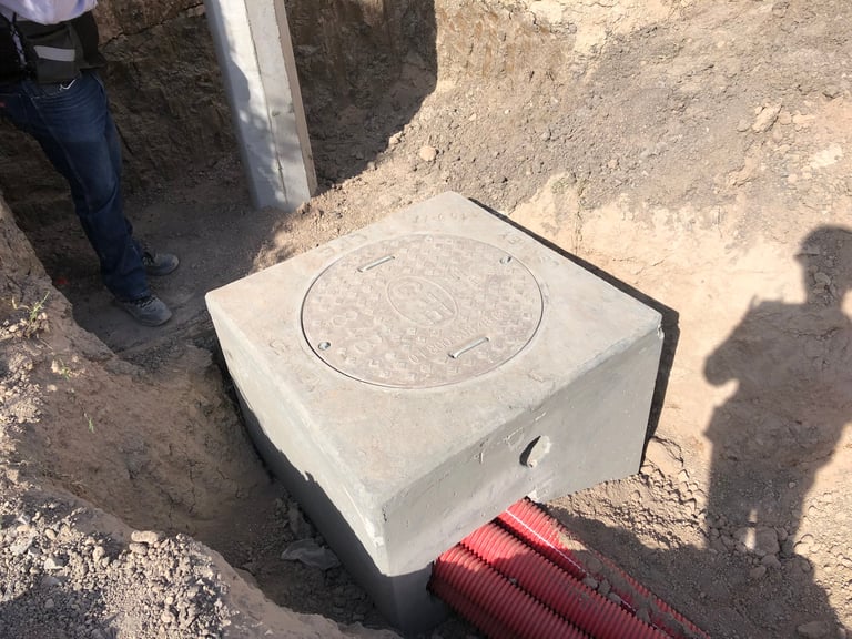 Concrete utility box with circular metal cover installed on ground next to red corrugated pipe and workers shadow