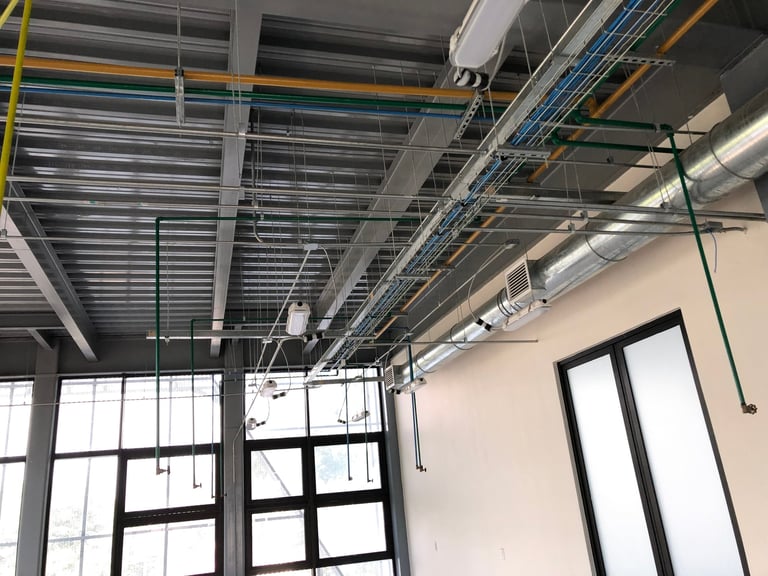 Industrial warehouse ceiling with exposed metal ducts, pipes, and corrugated panels, featuring large windows and bright overhead lighting