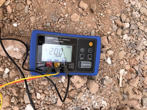 Digital multimeter with blue case displaying 2.00 on LCD screen, placed on sandy ground with test leads attached