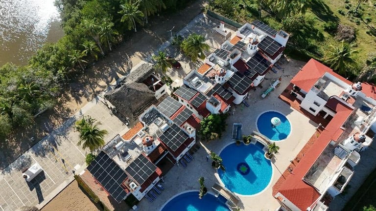 Aerial view of beachfront resort with multiple blue pools, red-tiled buildings, and solar panels surrounded by palm trees